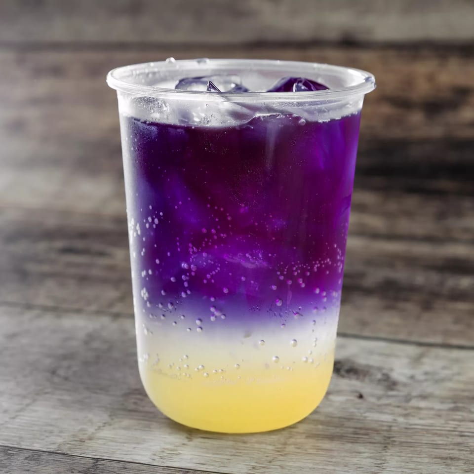 Layered purple and yellow sparkling beverage with ice in a clear plastic cup on a wooden table.