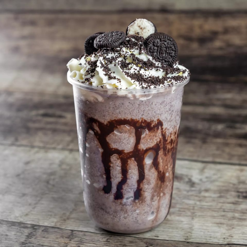 Chocolate milkshake in a clear cup with chocolate syrup, whipped cream, cookie crumbs, and chocolate sandwich cookies.