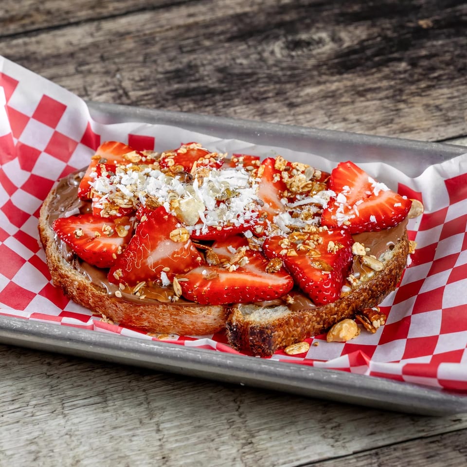 Toast topped with chocolate spread, sliced strawberries, granola, coconut, and nuts in a checkered-lined tray.