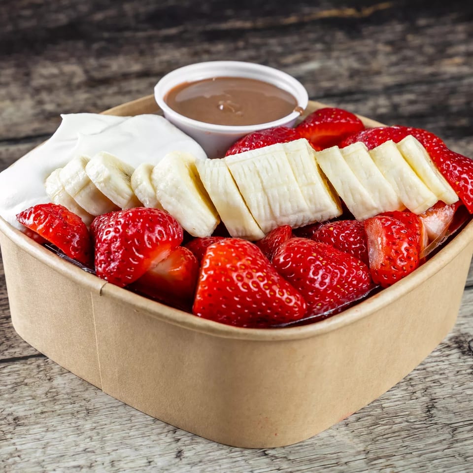 Bowl of sliced bananas, strawberries, whipped cream, and chocolate sauce on a rustic wooden table.