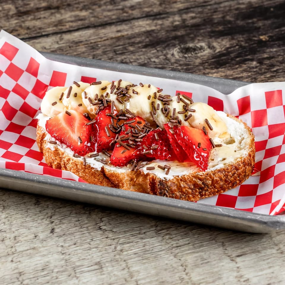 Thick toast topped with whipped cream, bananas, strawberries, red berry sauce, and chocolate sprinkles in a checkered-lined tray.